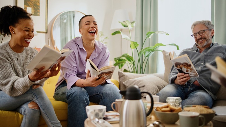 Three people laughing as they look at open books on a couch