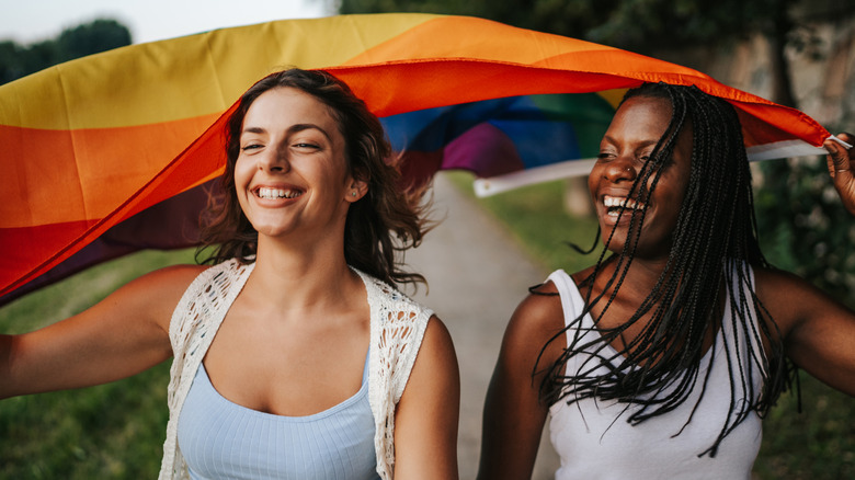 Two smiling women flying the gay pride flag behind them outdoors