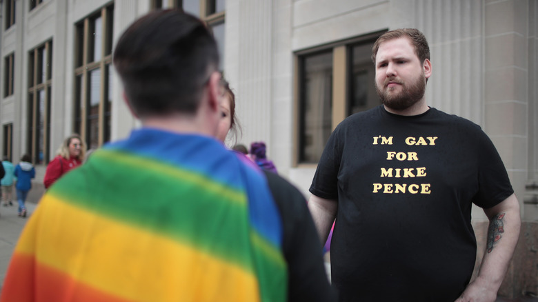 Man stands with others at a Pride event, one wearing a rainbow Pride flag
