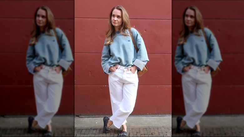 A woman wearing white jeans and a blue sweatshirt in front of a red wall